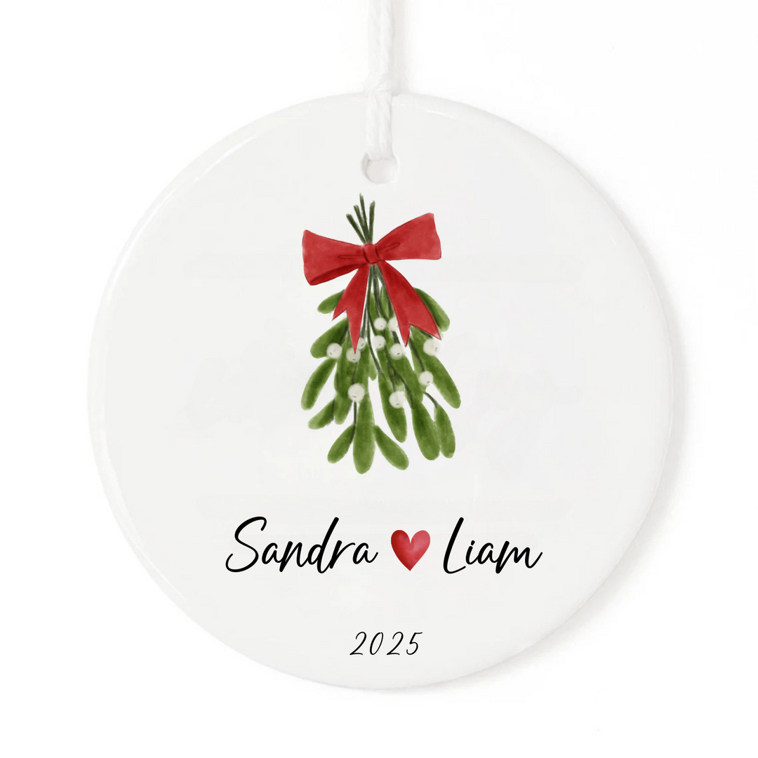 A white ornament with a mistletoe and red bow and a personalized couple's names and a date.
