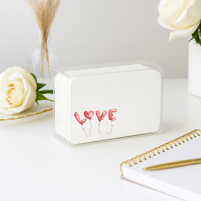 Small white box with 'LOVE' balloons design on a light surface with flowers and a notebook.