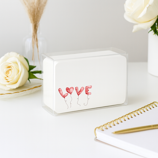 Small white box with 'LOVE' balloons design on a light surface with flowers and a notebook.