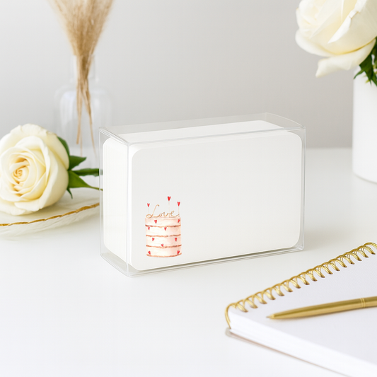 Clear cube with a cake design on a white surface with flowers and a notebook.